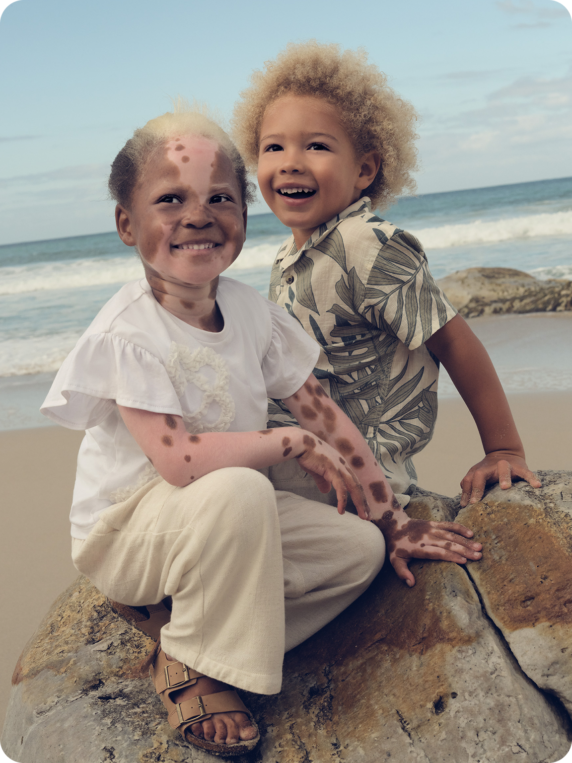 Two young models sat on the beach. Young girl wearing a linen white shirt and beige trousers. A young boy wearing a beige and green shirt. 